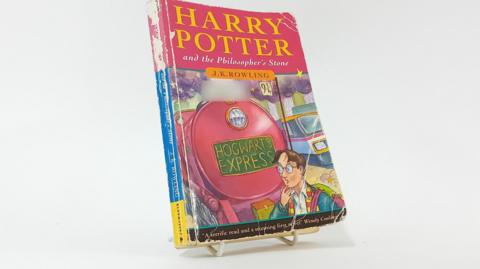 A paperback book which is worn at the edges and has a dog eared corner bears the words Harry Potter and the Philosopher's Stone. It also has a sticker saying 'I belong to' on the front. 