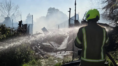 firefighter in a fluorescent helmet aims a hose into a smoked building where corrugated iron sheets and fire-damaged wreckage can be seen