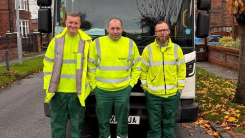 Three binmen wearing high-vis clothing, standing together and smiling in front of their lorry