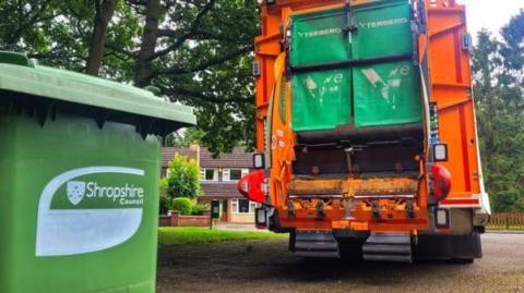 A green Shropshire Council bin in front of an orange bin lorry