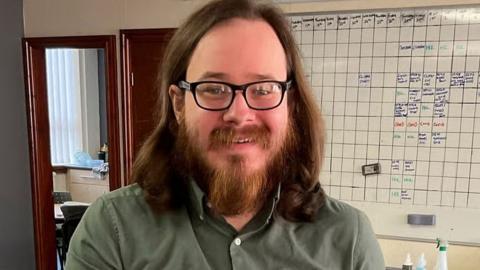 A photo of Joshua Kerry, a man with shoulder-length brown hair, a beard and glasses, from the shoulders up. He is smiling at the camera, wearing a khaki shirt, and behind him is a whiteboard.