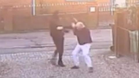 A person dressed in all black hitting a man, wearing white trousers and a dark top, with a wooden plank on the driveway of a house.