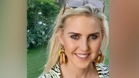 A smiling woman with long blonde hair, hoop earrings, and sunglasses resting on her head stands outdoors in front of green grass and flowering bushes. She wears a patterned outfit and look directly at the camera.