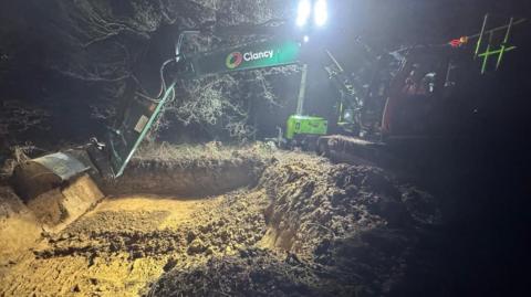A digger removing what appears to be sand from a wooded area at night