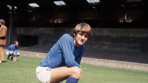 A young man with Beatles-esque dark brown hair wearing a blue football kit and crouching on a football pitch.