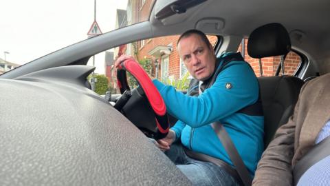 Chris Riley is sat in the driver's seat of a car, the camera is on the dashboard. His hands are on a red steering wheel and he is looking to the left as if checking the road is clear to enter. He has shaved black hair and wears a bright blue zipped up hoodie, with black detailing on the top half and blue jeans.