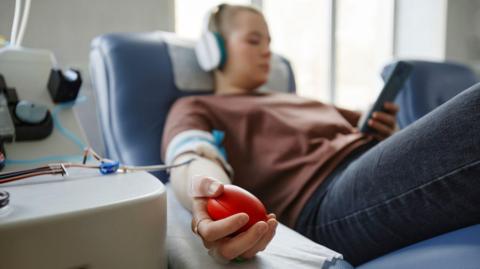 A stock photo of a woman sat in a blood donation chair. She's wearing dark blue jeans and a brown jumper, and has a pair of headphones in and a mobile phone in her left hand. Just her right hand is in focus - where she's squeezing a stress ball to keep up her blood pressure. Her right arm has plastic tubes coming out of it.