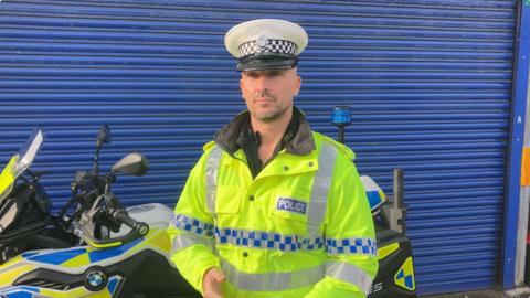 A man stands in front of a police bike and a blue shutter. He is wearing a flourescent yellow jacket with police emblazoned on the top pocket, and a white police hat