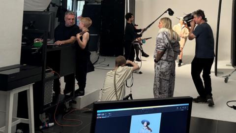 Six people taking part in a fashion shoot. Man on the far right is holding a camera. Woman stood next to him is wearing a grey patterned jumpsuit with long blonde hair. Kneeling on the floor is Liz wearing a cream top. Sitting at the computer to the left of the picture is Rankin wearing a black top and has a grey beard and hair talking to Liz's little brother who is standing on a white box and wearing black top and trousers. In the middle of the picture is a man in a black top holding a long-handled lamp and in the background is a woman wearing a cream coat. 