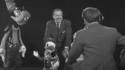 Walt Disney sits opposite interviewer Derek Hart, beside Donald Duck and Goofy figurines