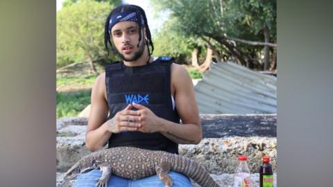 Ellis Heather has a blue headband on and is wearing a black, sleeveless stab vest. He has a komodo dragon on his lap and is sitting in Epping Forest.