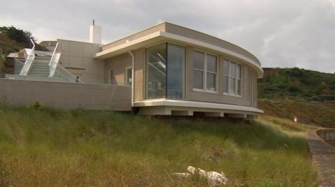 A picture of the site with a large grass verge in front of it. The building is cream and brown in colour.
