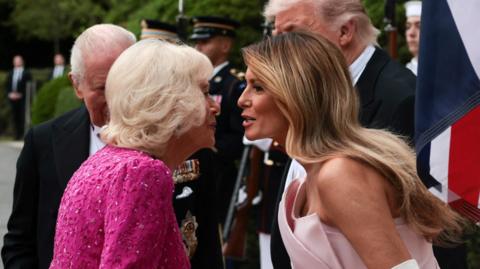 Queen Camilla wearing a dark pink dress and First Lady Melania Trump waring a pale pink dress go to kiss each other in greeting 