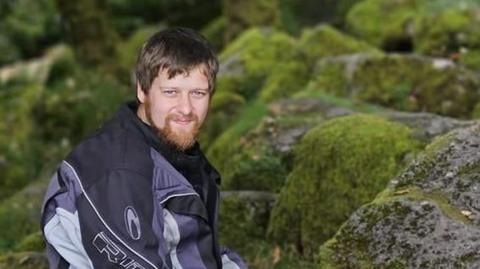 Ben Rowe, with a beard, is wearing a motorcycling jacket and sitting on rocks covered in moss in his Facebook profile picture