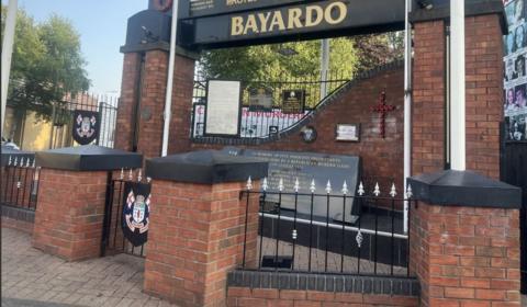 Memorial for those killed in the Bayardo Bar attack on the Shankill Road. There is a large sign saying Bayardo in black and gold at the top. There is also a black and gold memorial stone in the middle and railings at the front.