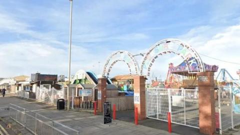 Entrance to a small seaside amusement park with white metal gates and brick pillars, decorative archways overhead, and rides visible inside under a bright sky.