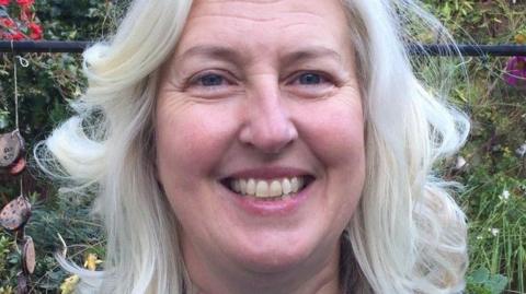 Headshot of Sarah Marriott. She has white hair and blue eyes and is smiling into the camera.  Behind her, foliage from a garden can be seen.