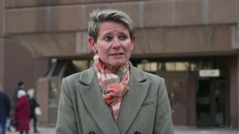 Cath Cummings, who has short hair with blonde highlights and wears a light grey-green mac coat with a pink and white patterned scarf, speaks to the camera with the brown façade of Liverpool Crown Court in the backdrop