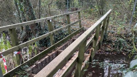 A small wooden bridge with metal fencing and trees on the far side and a small stream flowing underneath it