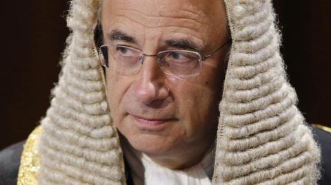 Sir Brian Leveson wearing a periwig in his former role as Lord Justice Leveson in 2015. His eyes look off to the side, framed by thin glasses. The background is dark.
