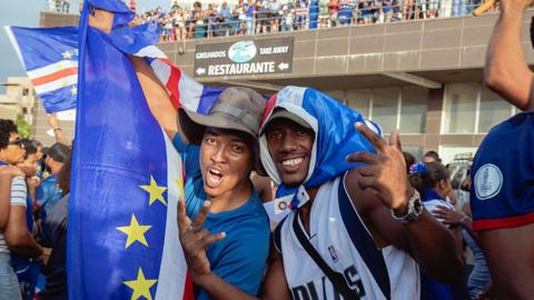 Supporters celebrate Cape Verde's victory. Two fans are draped in the country's flag and smiling at the camera.