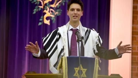 A Rabbi preaching in a synagogue, wearing a suit and purple tie and a black and white robe 