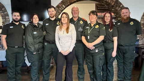 Sian Deeks, who has long, straight auburn hair and is wearing a grey V-necked top and black trousers, stands in front of seven paramedics - four men and three women - wearing dark green short-sleeved shirts and trousers.