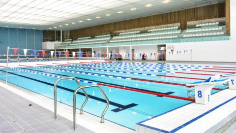 A large pool with multiple lanes and bunting hanging across it above the water. There are a number of seats in the stands which are empty.