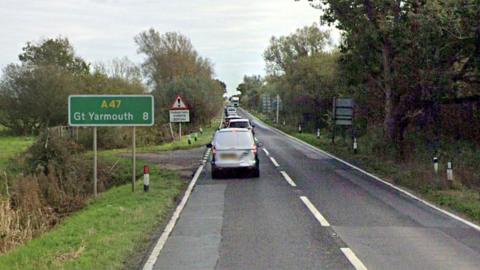 A stock image of the A47 Acle Straight showing cars backing up. The road is one lane in either directions. A road sign showing Great Yarmouth is on the side of the road, which is surrounded by fields and shrubbery.