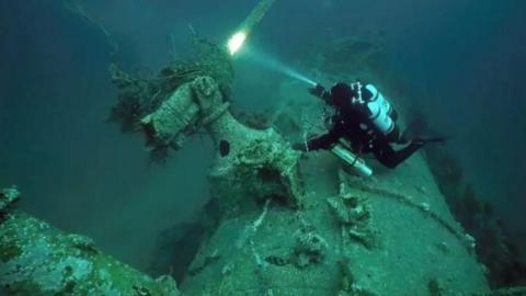 The image is taken underwater and shows the German U-boat on the ocean floor. There is algae across a lot of the vessel. A diver can be seen in a black wetsuit above the vessel. They are shining a torch on a part of the U-boat.