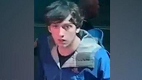 A CCTV shot of Thomas Dures, 21, who is wearing a blue coat and a black t-shirt and has medium-length dark hair