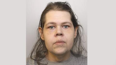 A police mugshot of Christine Kekkonen. She is wearing a grey t-shirt and standing against a light-grey background. She has grey-blue eyes and brown straggly hair. She has a blank expression on her face and is staring into the camera.