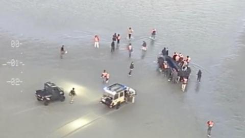 Two cars in the shallow water with people surrounding what appears to be a dingy boat with people inside the vessel