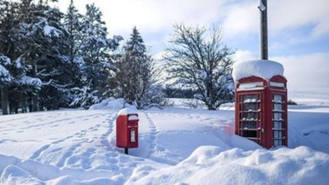 A snow covered red post box and phone box surrounded by deep snow and trees behind