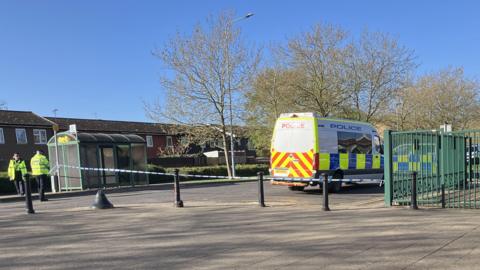 A crime scene near a bus stop. Two officers in high-vis vests are standing in the road, and a police van and car are parked on the street. Police tape is in place.