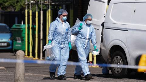 Two people wearing forensic protective suits, hair coverings and gloves walk beside a white van behind a police cordon. Bollards and tape are visible in the foreground, with a road with cars and bins in the background