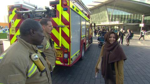 A woman wearing a brown headscarf smiles as she talks to two LFB firefighters in uniform next to a red and yellow striped fire engine. The entrance to Stratford Station can be seen in the background