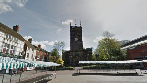 A town centre with a church in the centre of the image and market stalls in the foreground. There are pubs and shops surrounding an open public square.