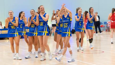 Team Bath Netball squad. A group of women all dressed in royal blue kit with yellow detailing and white socks with white trainers are standing on the indoor netball court. They are all smiling and clapping, with some looking at the camera while others look at each other or out to the crowd. 