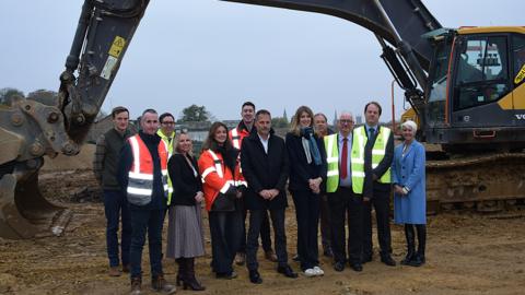 A group of 12 men and women, some wearing hi-vis vests and jackets, stand for a photograph to celebrate the next stage of the project. There are diggers in the background and they are standing on a piece of muddy land.