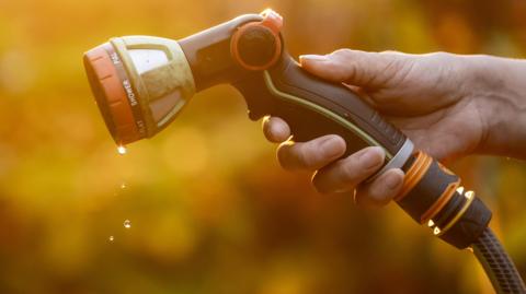 Close-up of a hand holding a garden hose spray nozzle, with water droplets falling from the nozzle against a blurred golden background.
