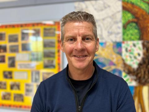 A man wearing a navy blue sweatshirt with a black zip. The man has grey hair and is standing infront of an art display