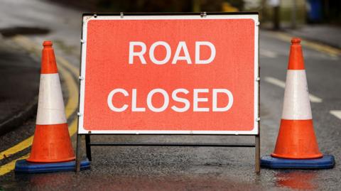 A rectangular red road sign stands on a road between two red and white cones. On the sign is printed "Road Closed" in white text.