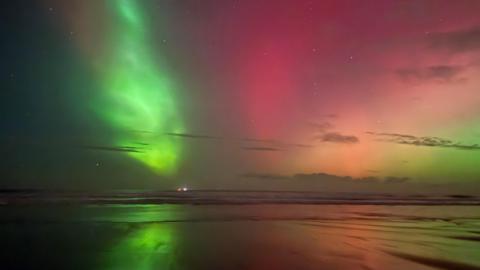 Green, pink, orange and pale yellow light glows above the beach and is reflected in the sea and wet sand.