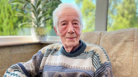 90-year-old Ernest Williams sat in a conservatory. There are large windows behind him showing blue sky and trees outside. 