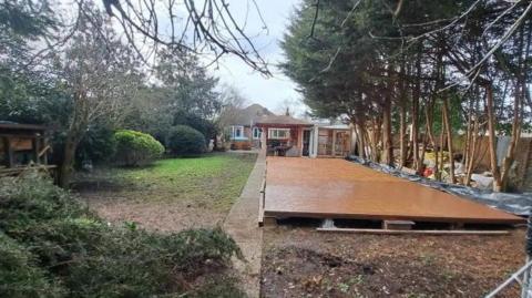 A residential property with a large wooden panel laid on the ground in the front garden of the house. There are trees to the right of the panelling.