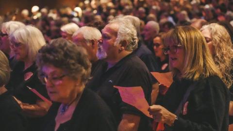 A view of hundreds of singers with a man and several women in the foreground