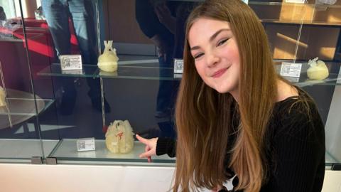 Jasmine in a black top and blue jeans pointing at the rubber replica of her heart that is behind a glass cabinet.