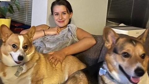 Hannah is smiling towards the camera with two corgis. They are all sitting on a dark grey sofa. Hannah has a light grey blanket on.