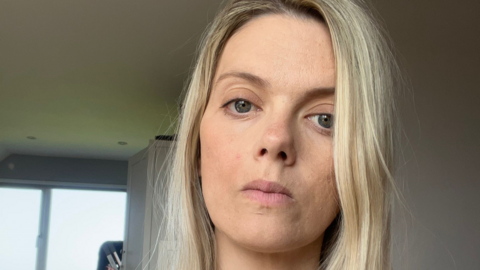 A selfie of Elen Hughes inside a house with light grey walls and a window visible behind her. She looks serious and has long blonde hair and there is a light grey cupboard behind her